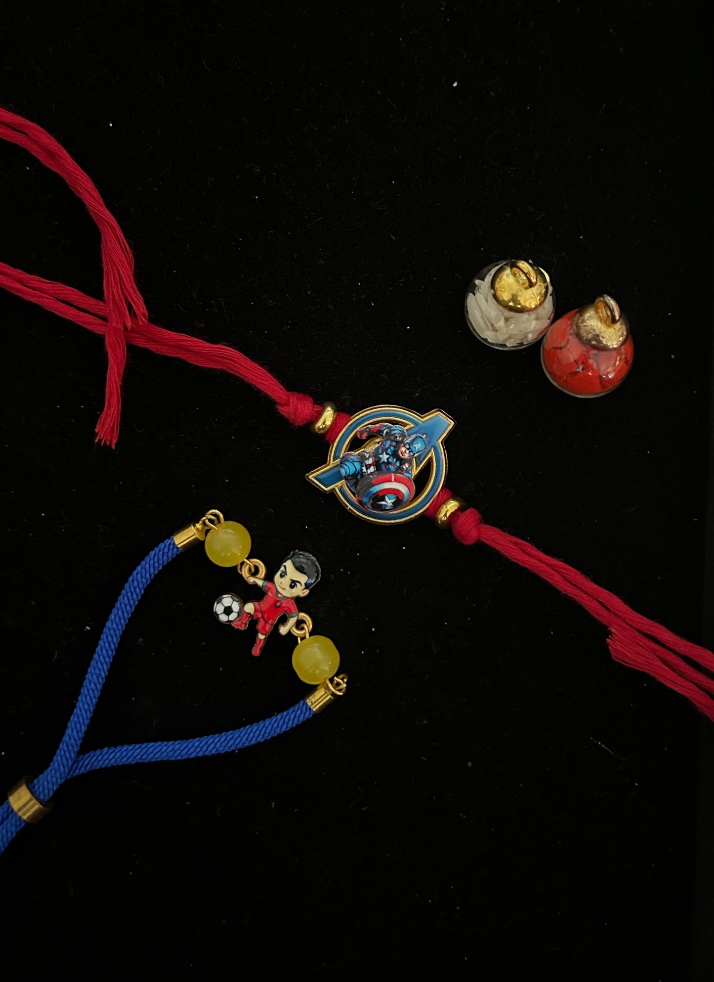 Two traditional Indian Rakhi string bracelets displayed on a black background. One Rakhi is red with a central circular charm featuring Marvel characters (Captain America and Black Widow) and small gold beads.