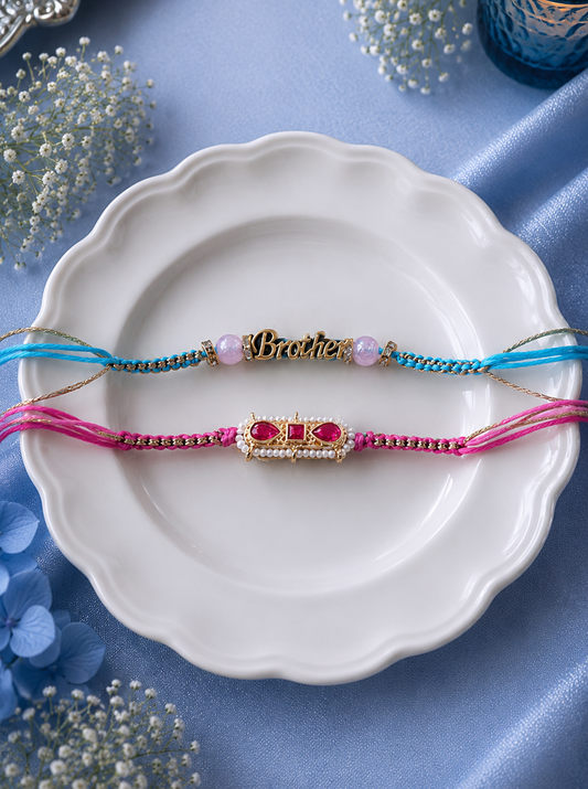 Two braided Rakhi bracelets, one in blue and one in pink, laid flat on a white scalloped plate. The blue Rakhi has a central gold-toned 'Brother' nameplate with a pearl, and the pink Rakhi has a gold-toned 'Brother' nameplate with red stones.